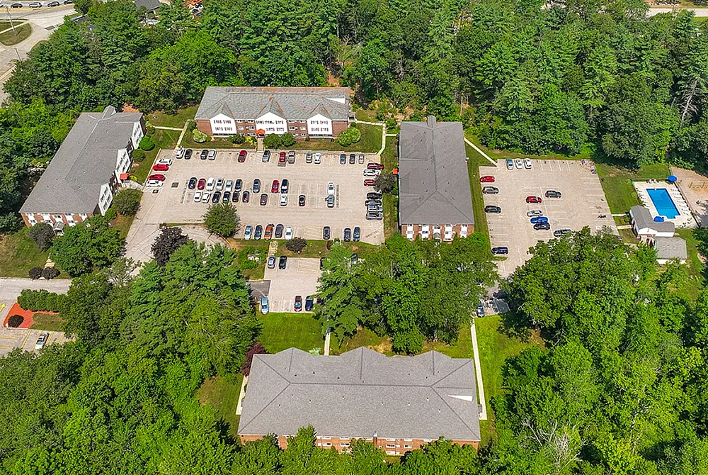 Aerial view of a scenic apartment complex nestled among lush greenery, featuring expansive parking lots and a sparkling swimming pool.