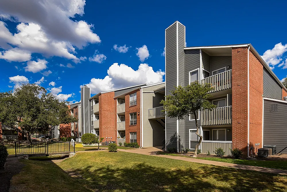 Charming apartment complex with modern architecture, featuring red brick and gray paneling, set against a vibrant blue sky with fluffy clouds.