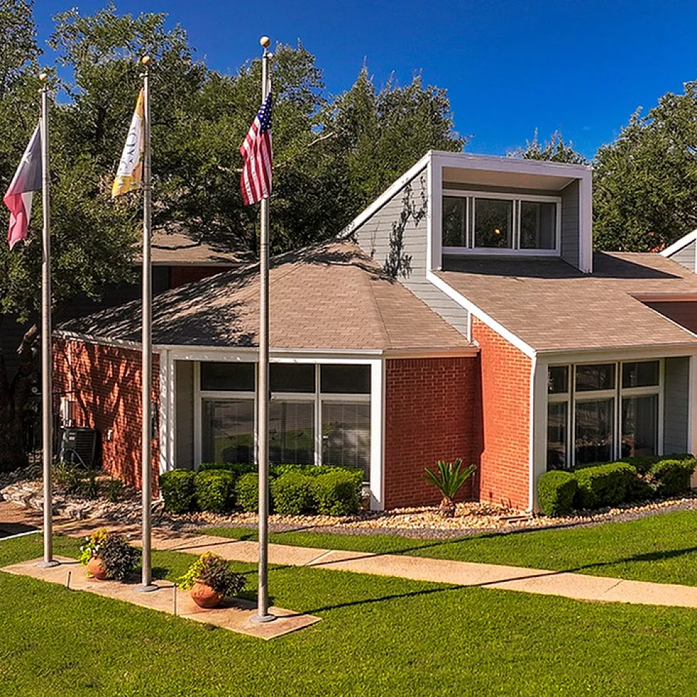 Charming brick community building with large windows, surrounded by lush greenery and three flagpoles, set under a clear blue sky.