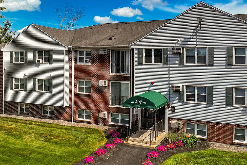 Charming apartment building with classic brick and siding exterior, featuring well-maintained landscaping and vibrant flower beds under a bright blue sky.