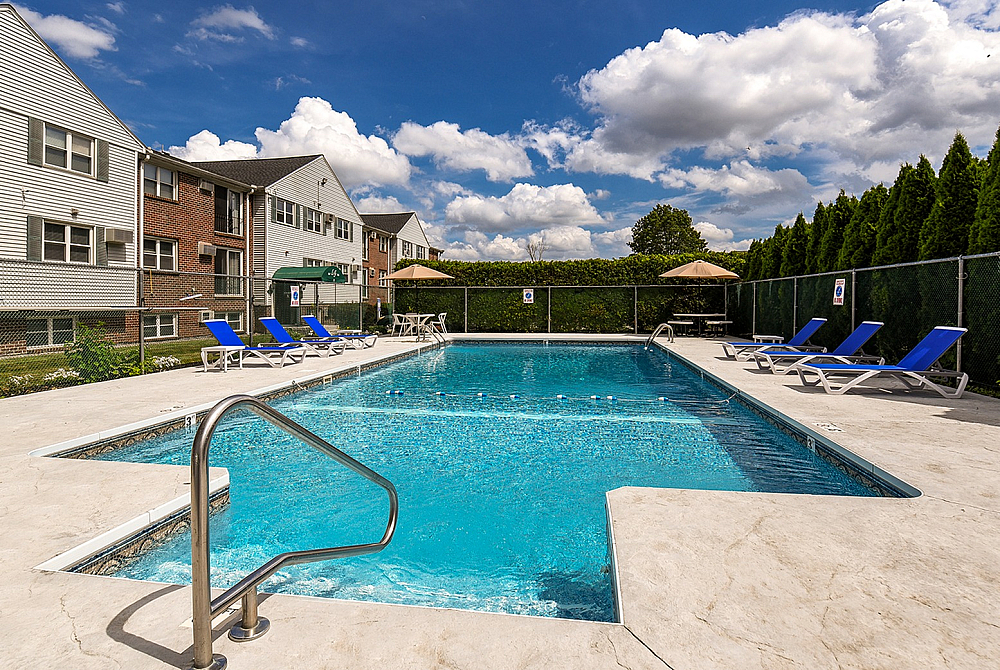 Inviting outdoor pool area with comfortable lounge chairs, surrounded by lush greenery and charming residential buildings under a sunny blue sky.