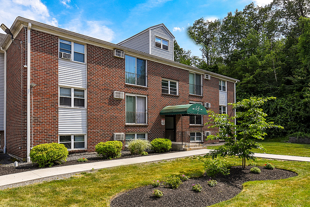 Charming brick apartment building with lush landscaping and a welcoming green awning entrance, surrounded by vibrant greenery and a manicured lawn.