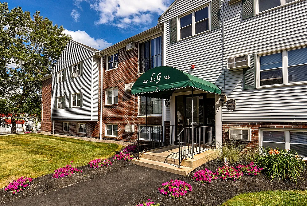 Charming apartment building with classic brick and siding facade, featuring a welcoming green canopy entrance and vibrant landscaped flowers.
