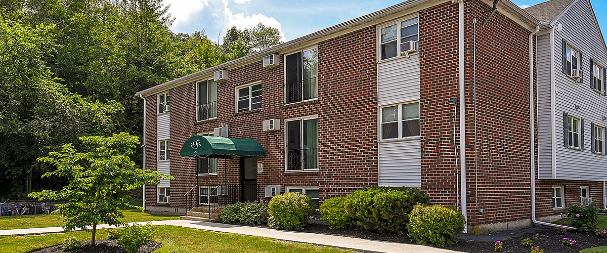 Charming brick apartment building with lush greenery and a welcoming entrance, perfect for comfortable and stylish living.