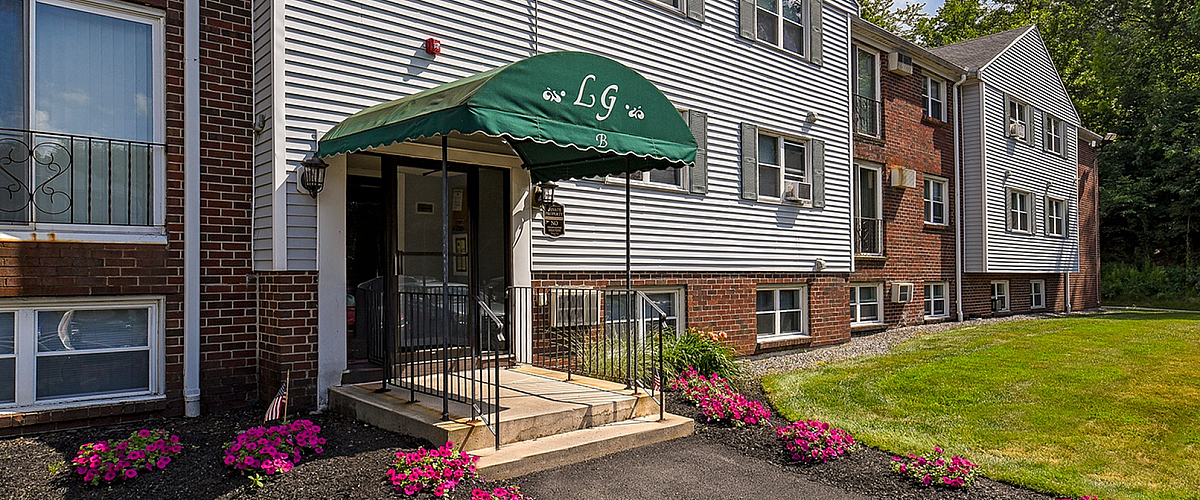 Charming apartment building entrance with inviting green awning, beautifully landscaped with vibrant flowers, set against a clear blue sky.