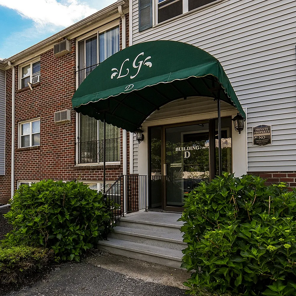 Charming residential building with a welcoming green canopy entrance, surrounded by lush greenery under a bright blue sky.