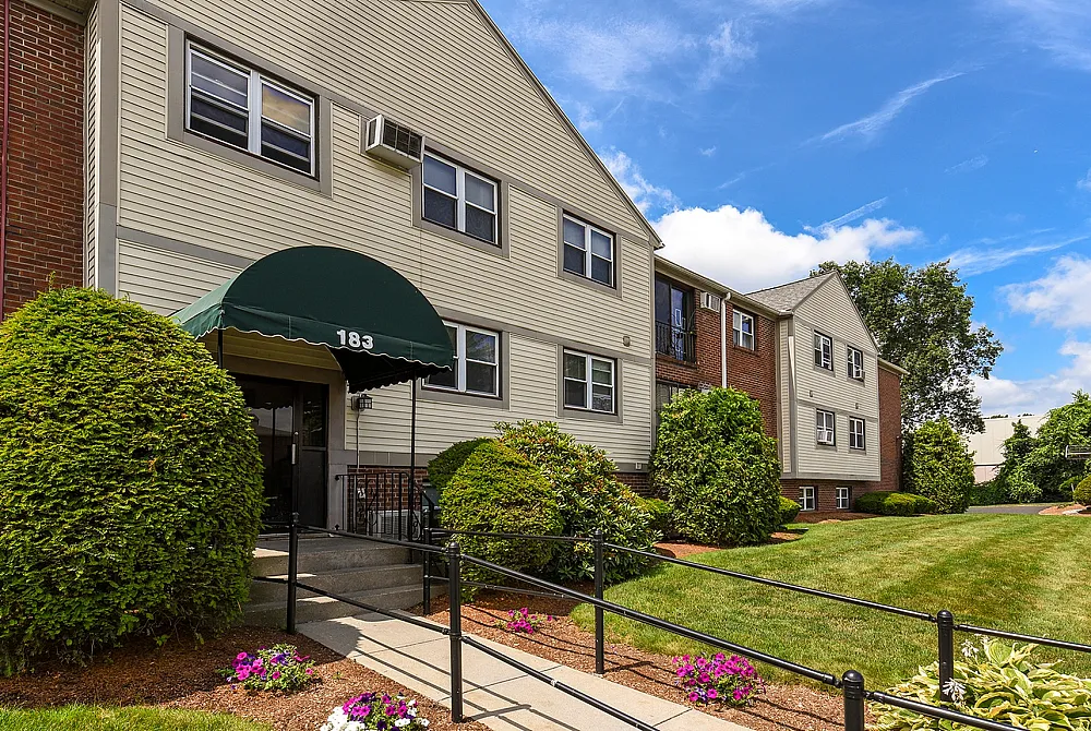 Charming apartment building with a welcoming green awning entrance, surrounded by lush landscaping and vibrant flowers under a bright blue sky.
