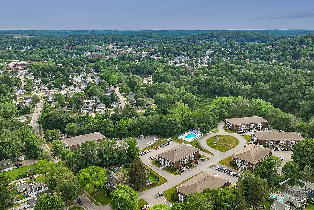 Scenic aerial view of a quaint residential neighborhood surrounded by lush greenery, featuring multiple apartment complexes with easy access to nearby amenities and a sparkling communal pool.