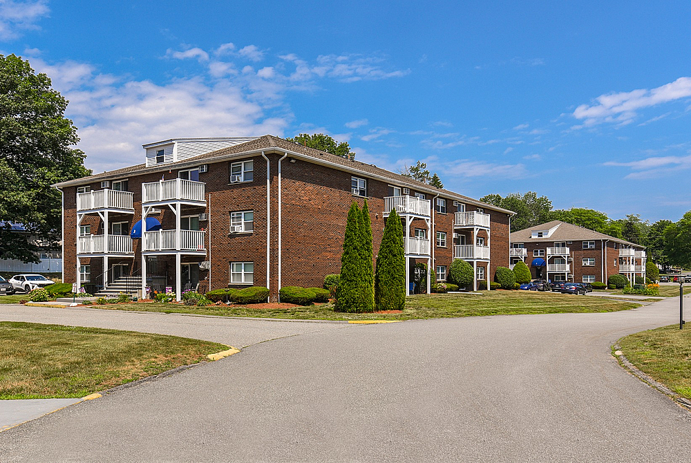 Charming brick apartment complex with spacious balconies, lush greenery, and ample parking under a clear blue sky.