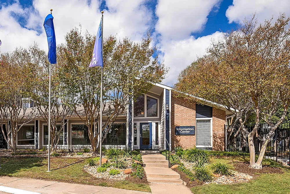 Inviting apartment information center with modern architecture, surrounded by lush greenery and flag poles, under a vibrant blue sky.