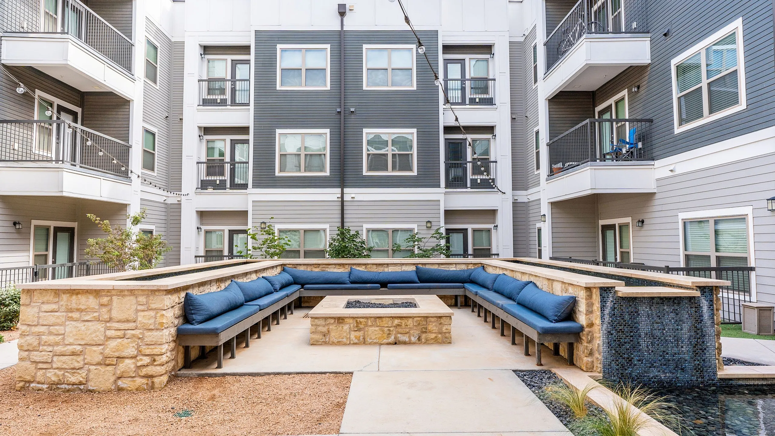 Modern apartment courtyard featuring elegant seating around a central fire pit, surrounded by contemporary multi-story buildings with balconies.
