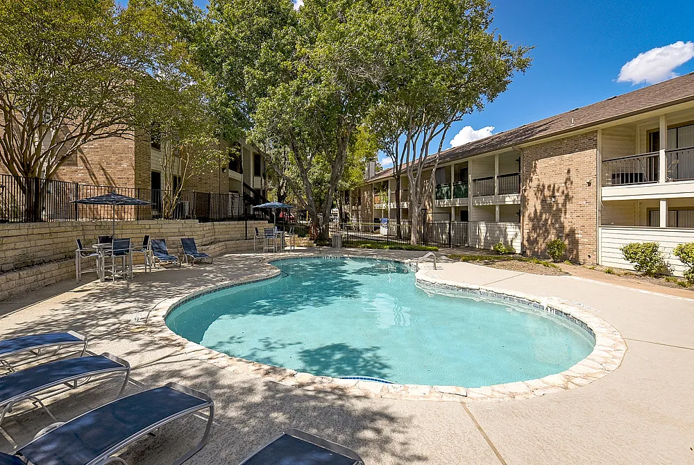 Charming apartment complex with a serene curved swimming pool, shaded by lush trees and surrounded by comfortable lounge chairs.