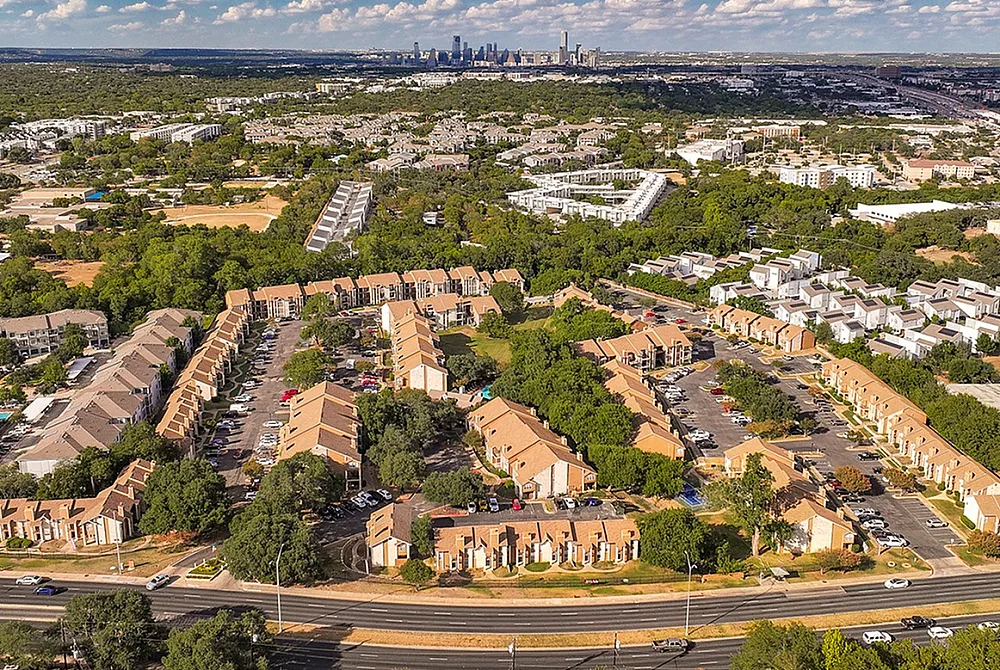 Aerial view of a vibrant apartment community surrounded by lush greenery and city skyline in the background.