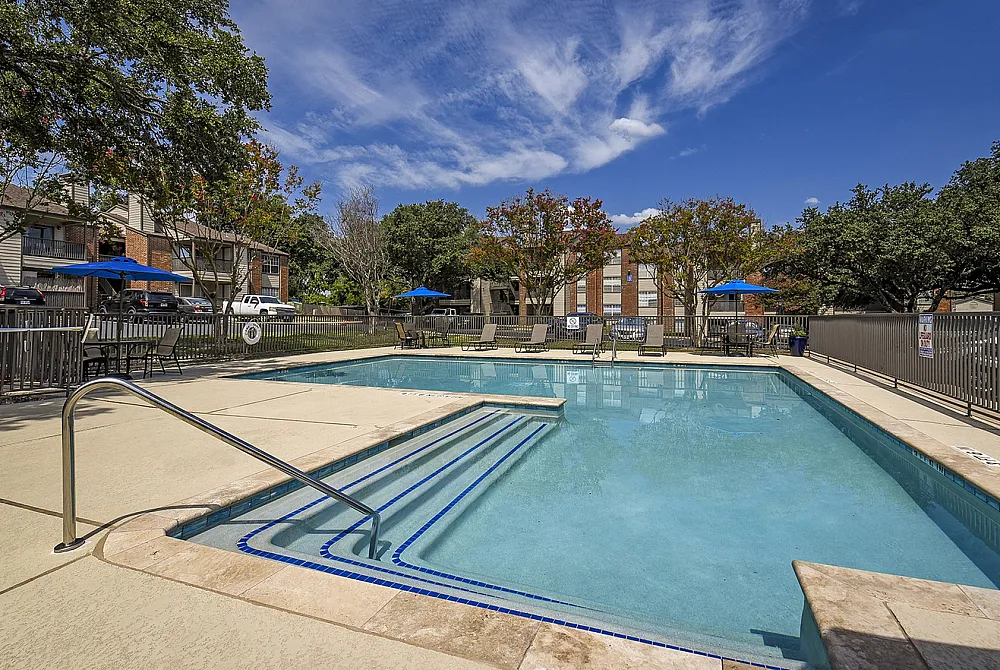 Inviting outdoor swimming pool area with surrounding lounge chairs and vibrant blue umbrellas, set against picturesque garden landscaping and clear skies.
