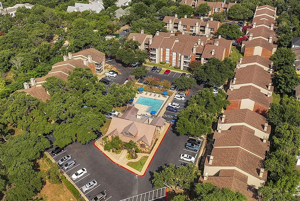Aerial view of a picturesque apartment complex with a central pool area, surrounded by lush greenery and neatly arranged buildings, offering a serene living environment.