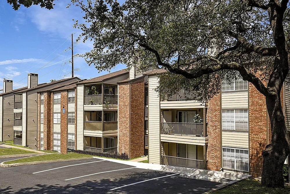 Charming apartment complex with brick facade and spacious balconies, surrounded by lush trees and ample parking space.