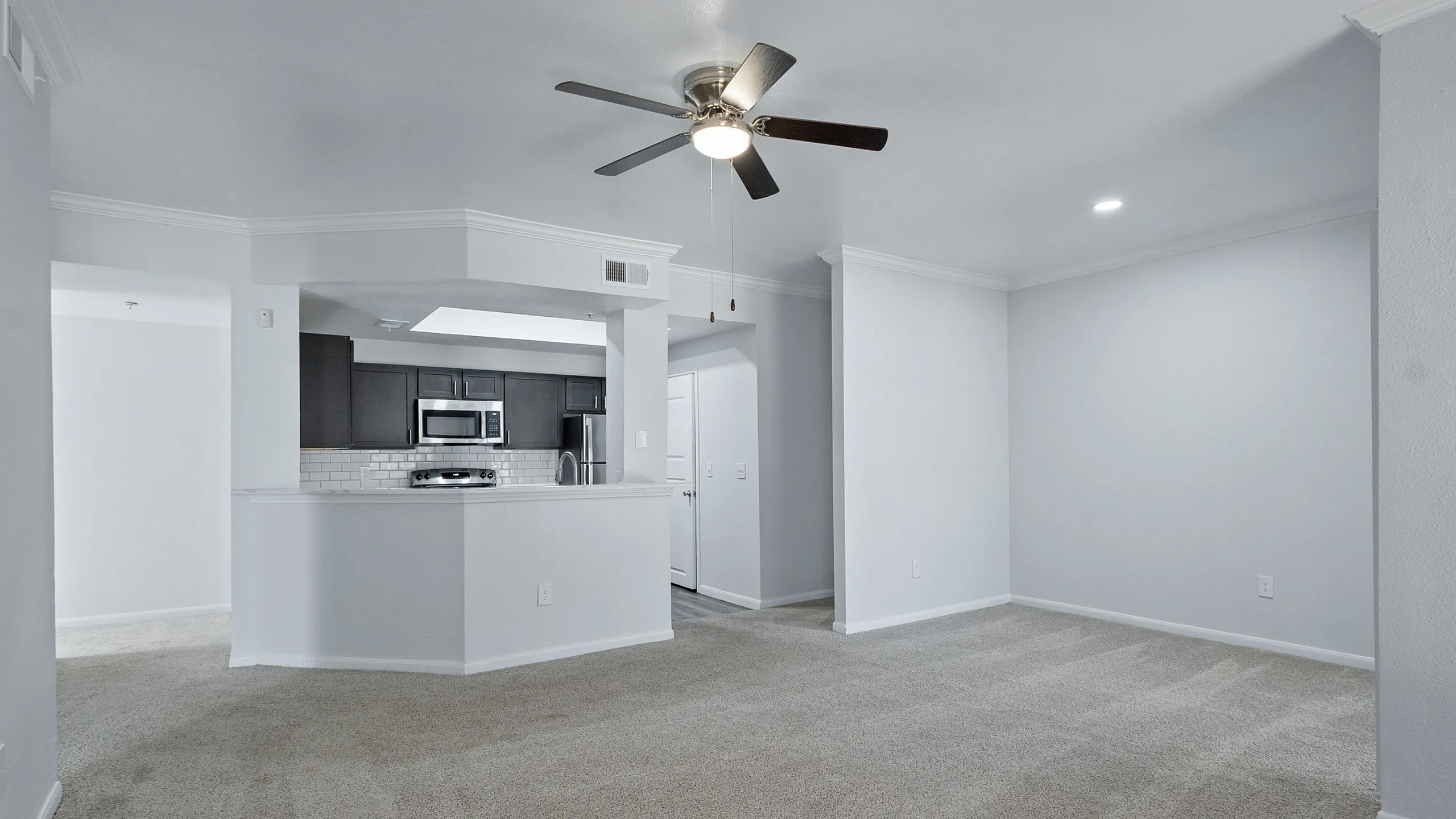 Bright and airy living area with plush carpeting, ceiling fan, and open-access kitchen showcasing modern stainless steel appliances.