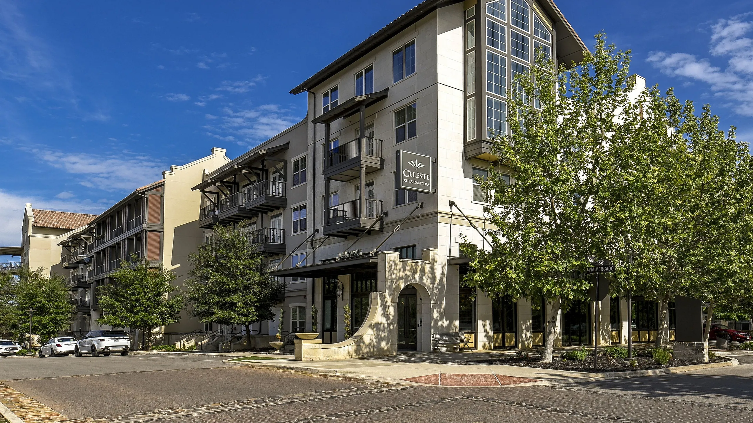 Elegant apartment building with modern design and balconies, surrounded by lush trees under a clear blue sky.