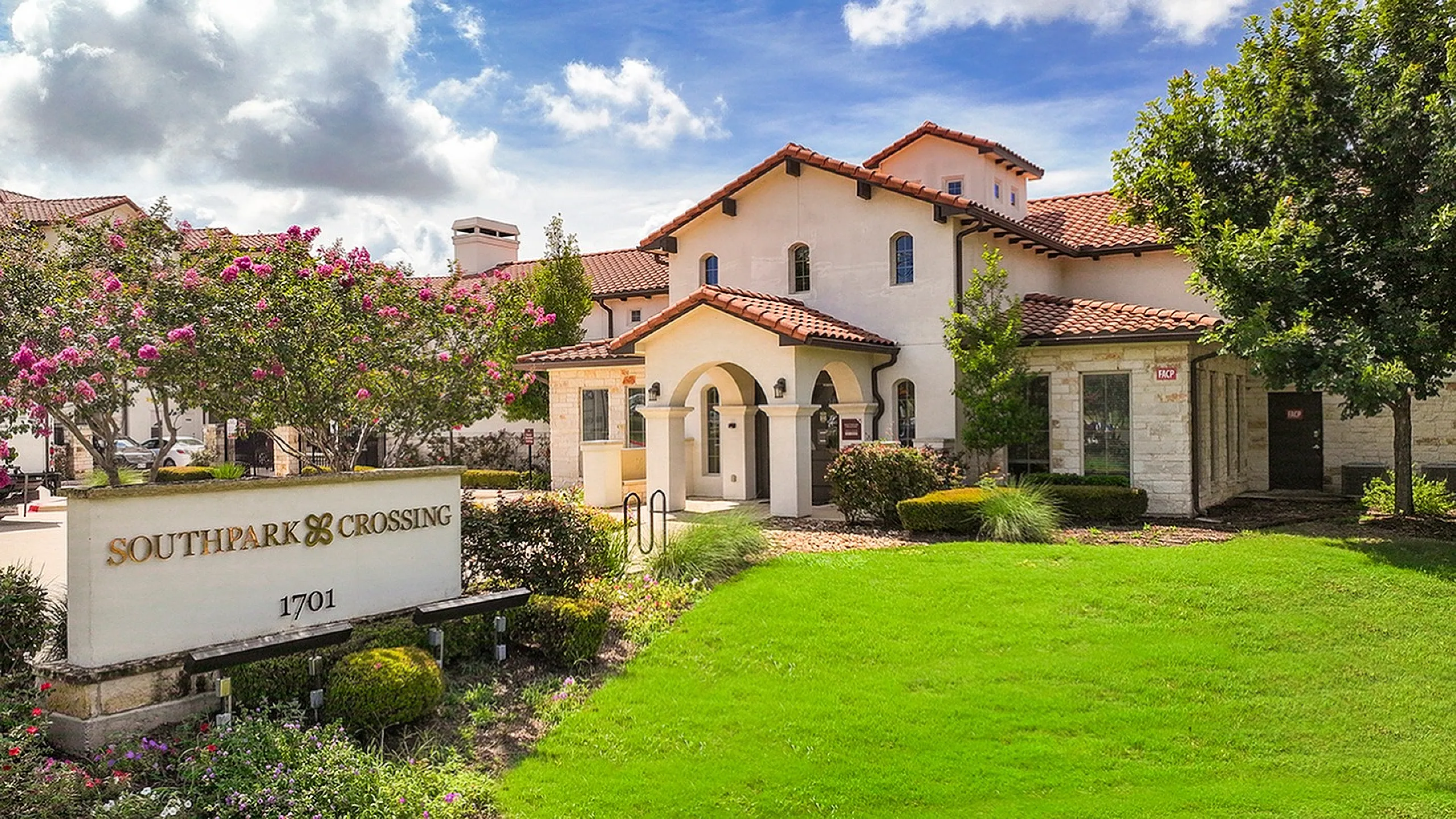 Charming Mediterranean-style apartment complex with lush landscaping and colorful flowering trees under a bright blue sky.