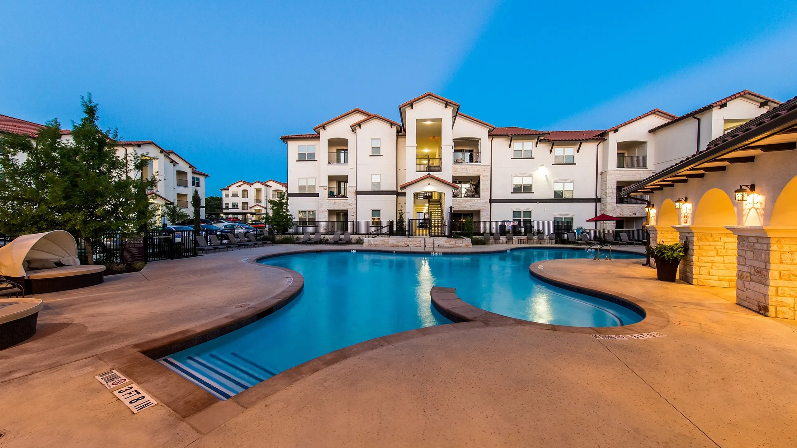 Luxurious apartment complex at dusk featuring a beautifully illuminated outdoor swimming pool surrounded by chic modern architecture.