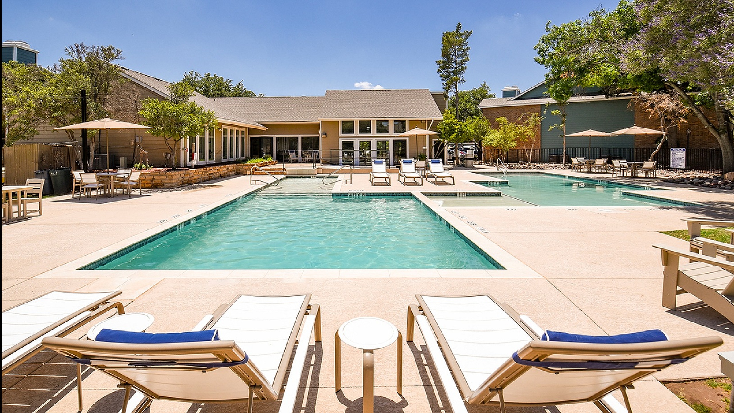 Inviting poolside oasis with modern lounge chairs, adjacent to a stylish clubhouse surrounded by lush greenery and clear blue skies.