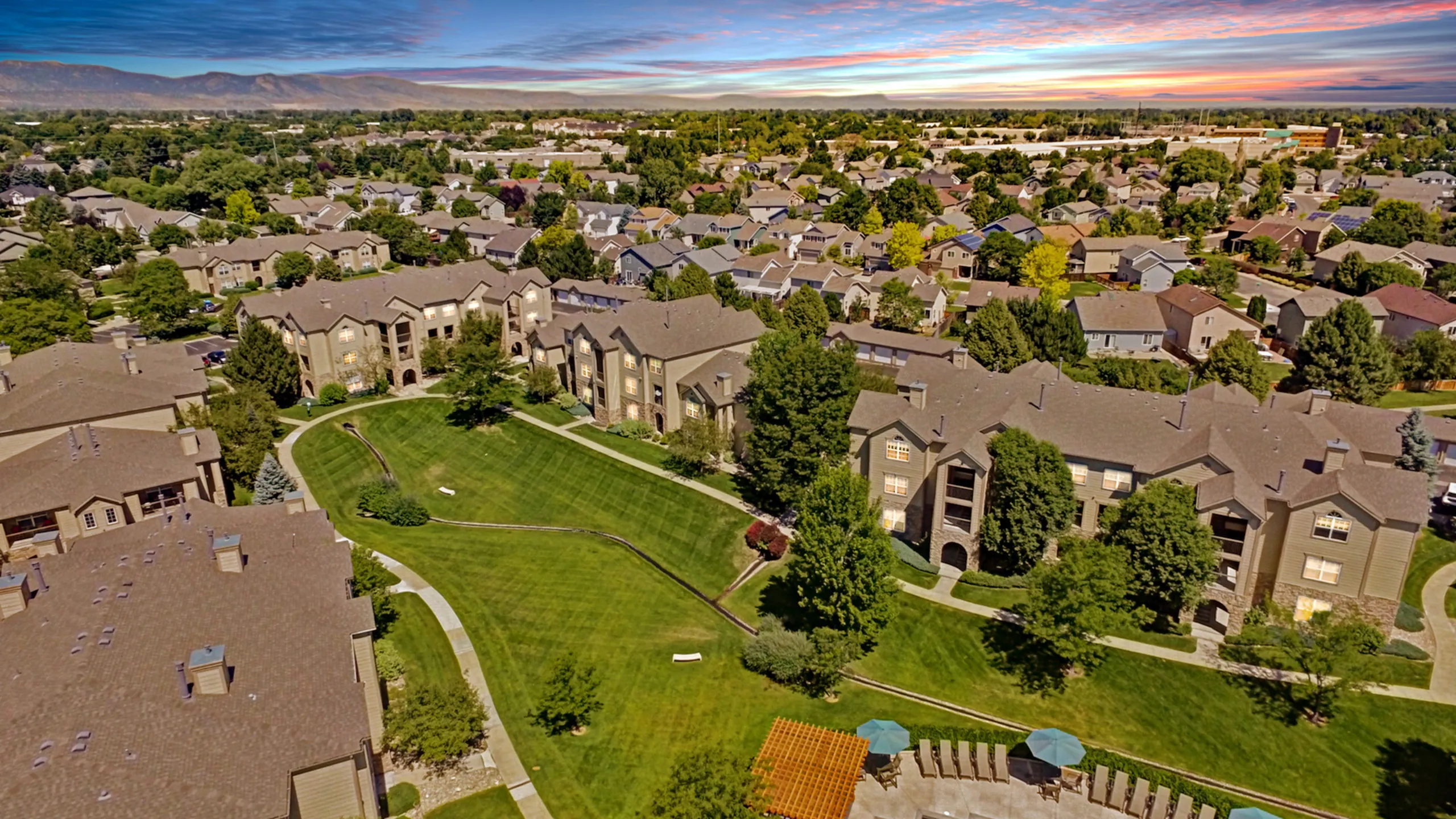 Aerial view of a tranquil apartment complex surrounded by greenery and residential homes under a vibrant sunset sky.