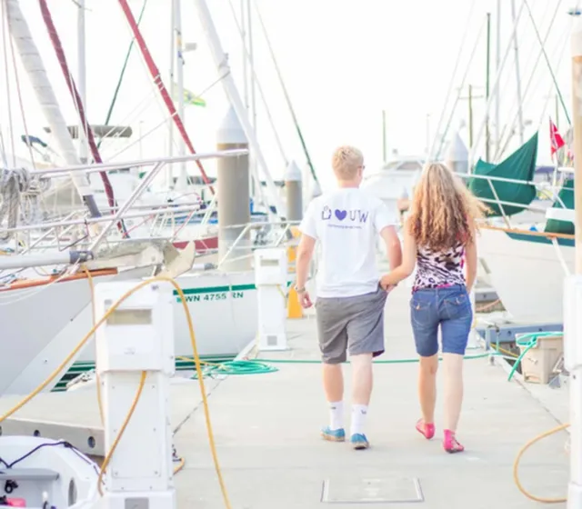 Couple walking hand in hand along a marina dock lined with sailboats.