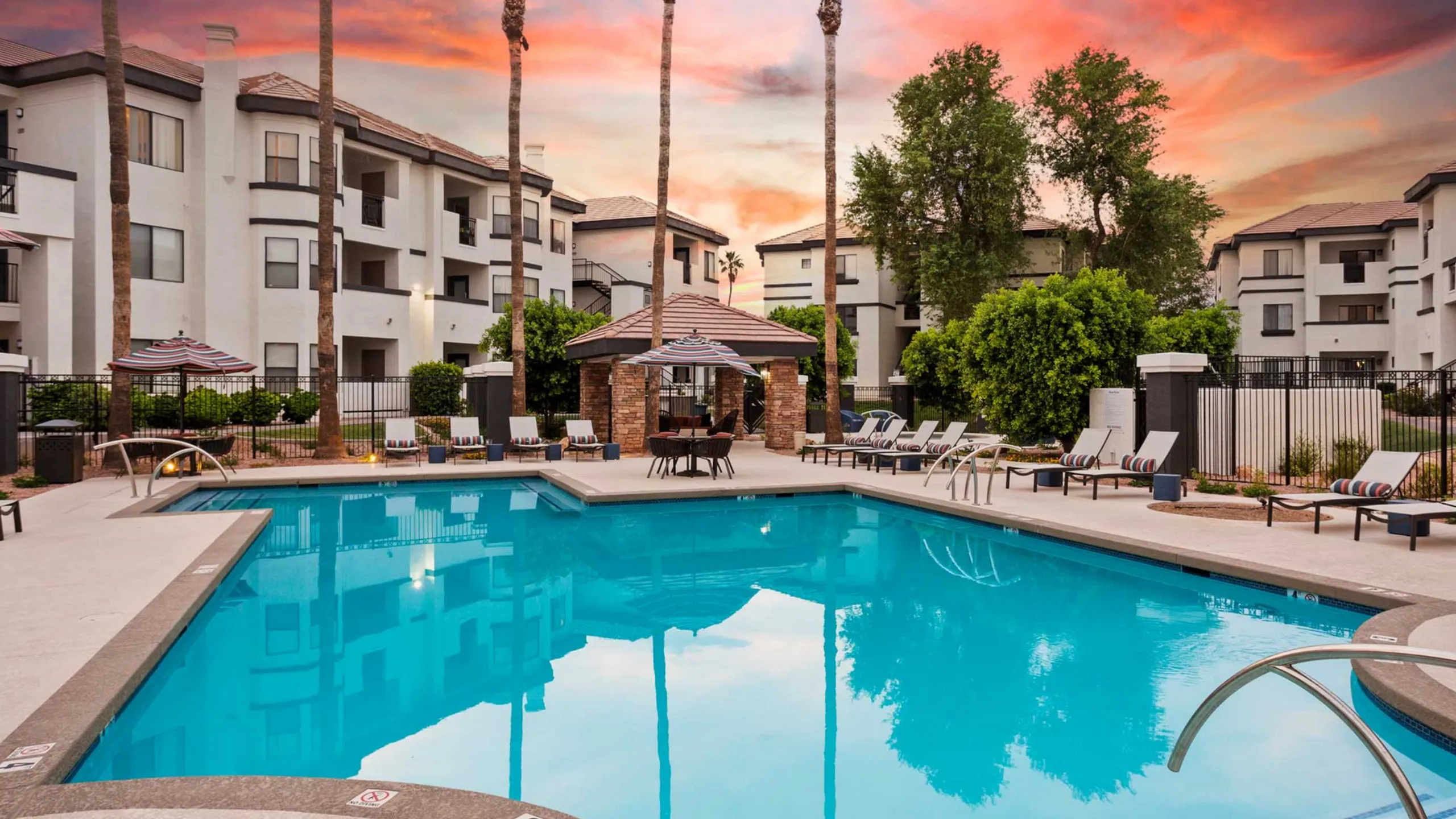 Luxury apartment complex with a shimmering pool, surrounded by palm trees and modern white buildings under a vibrant sunset sky.