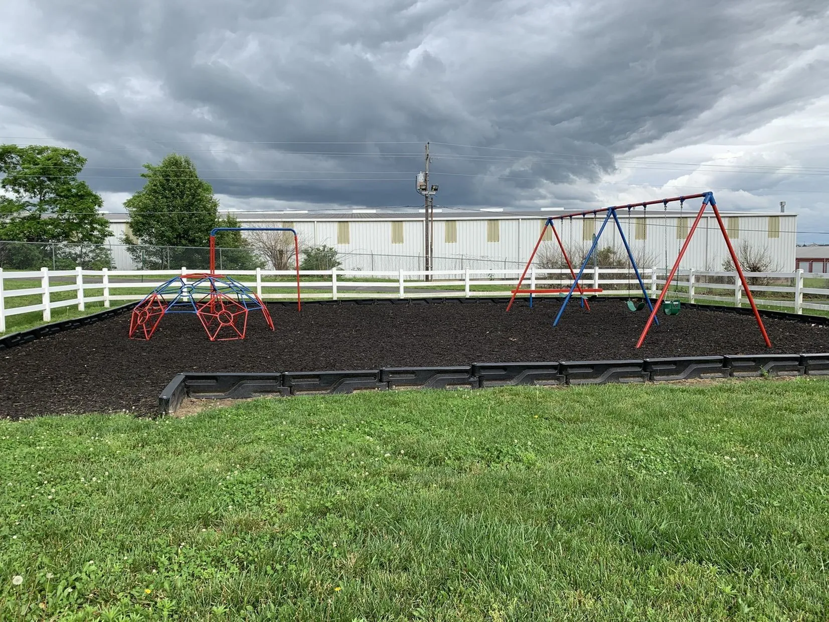 Charming playground with swings and a vibrant climbing dome, surrounded by lush greenery and a quaint white picket fence, ideal for family-friendly communities.