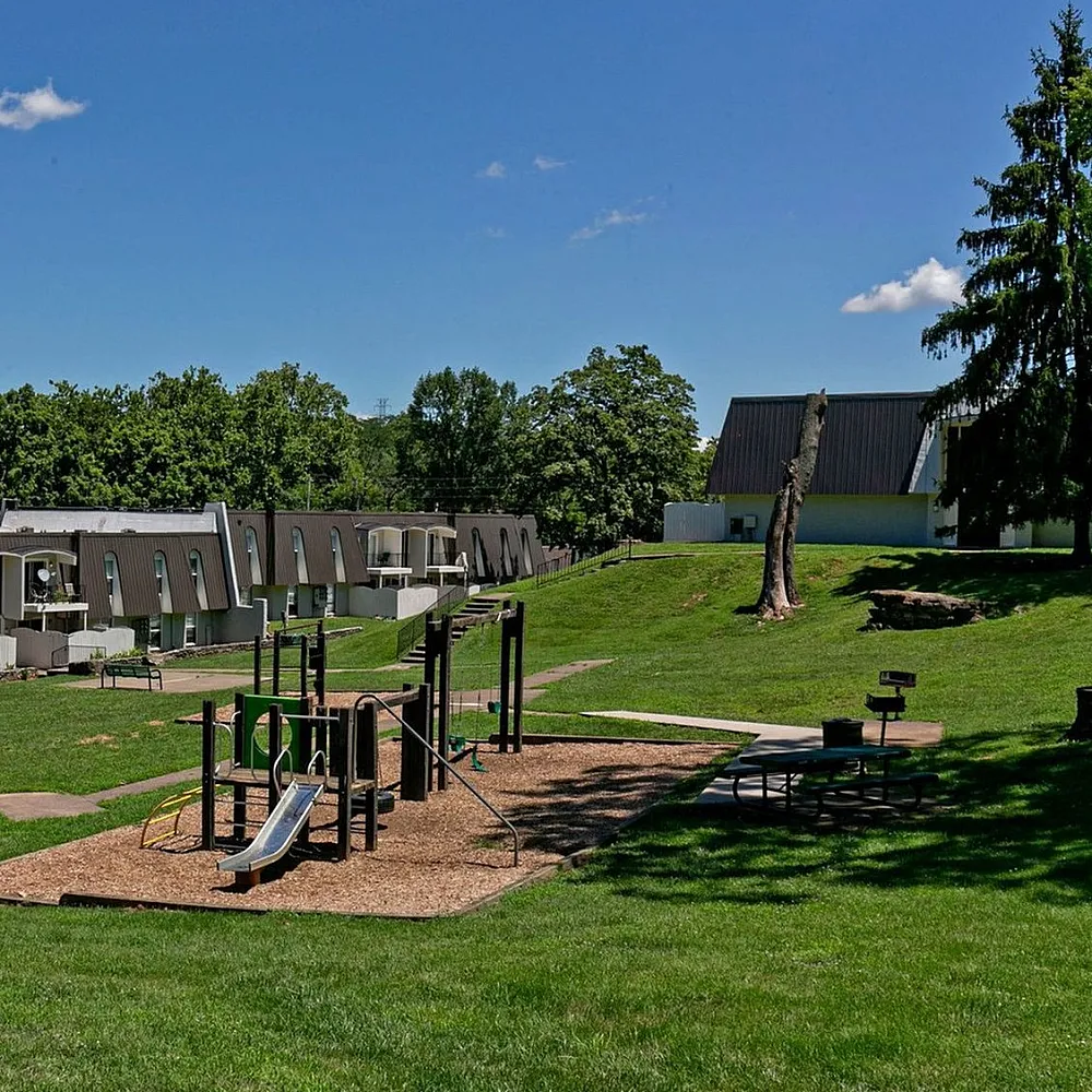 Charming residential complex with modern architecture surrounded by lush greenery, featuring a playground and recreational area under a clear blue sky.