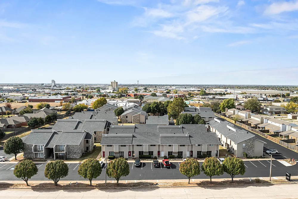Aerial view of a modern apartment complex with clean architecture, ample parking, and lush trees under a clear blue sky, offering a glimpse of city life in the distance.