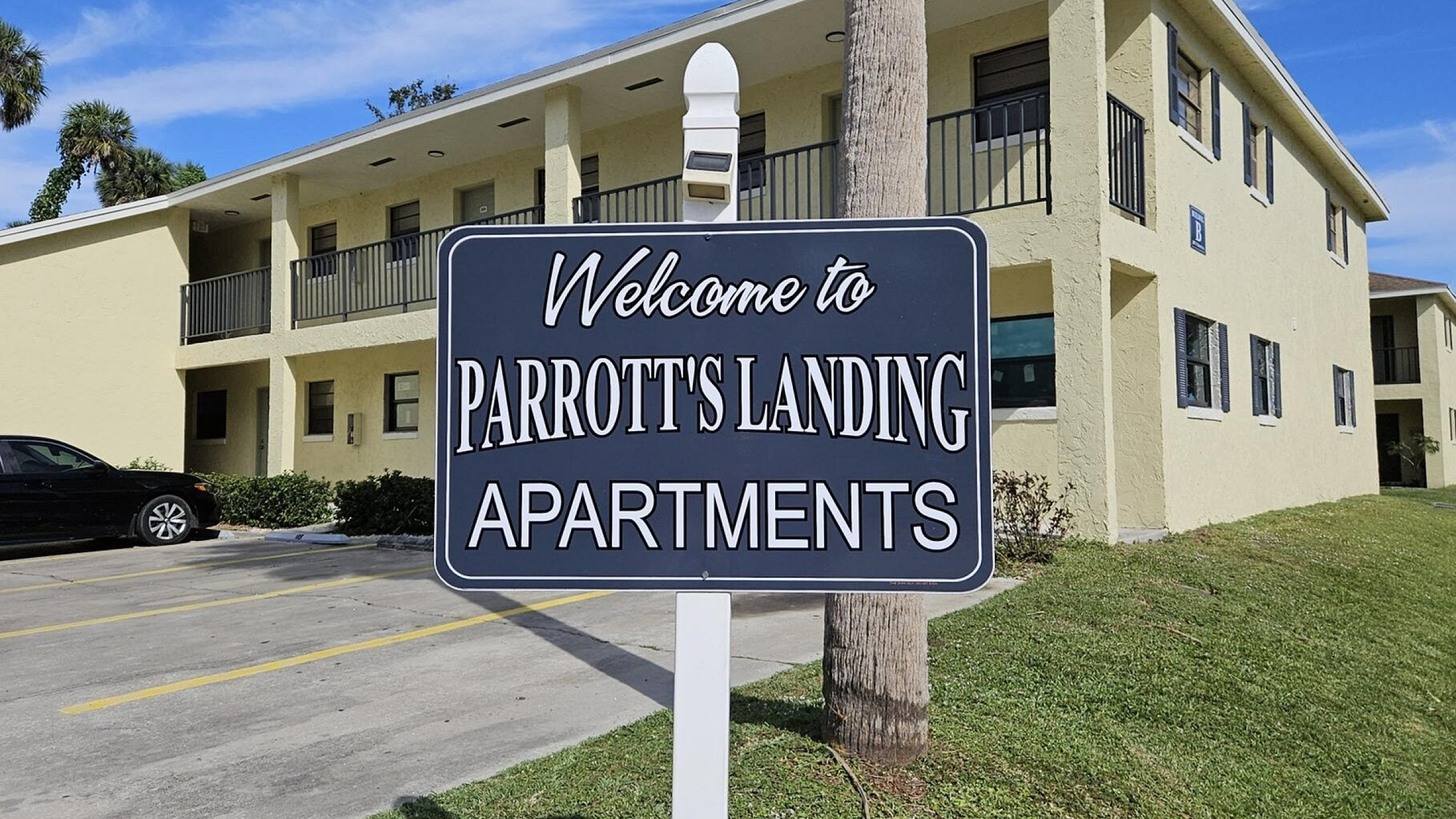 Inviting entrance to Parrott's Landing Apartments with lush greenery and modern architecture under a bright blue sky.