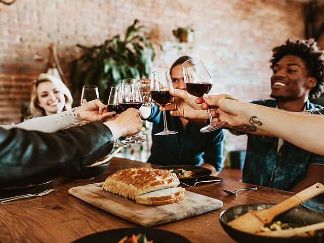 Lively group of friends toasting with wine in a stylish, rustic dining setting featuring exposed brick walls and a wooden table with fresh bread.