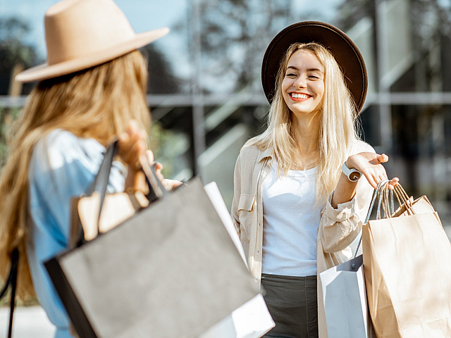 Two stylish women enjoying a sunny shopping day, smiling and carrying shopping bags, wearing trendy hats.
