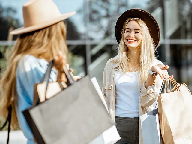Joyful scene of two stylish women wearing hats, laughing and holding shopping bags in an urban setting.
