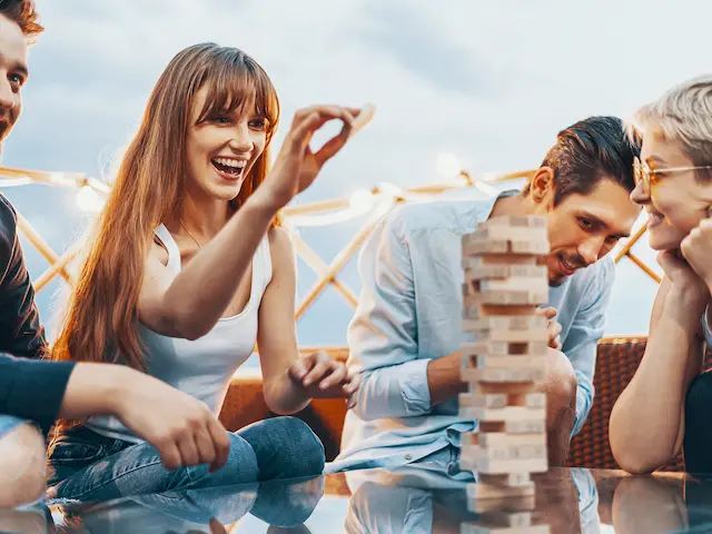Group of friends laughing and playing a game of Jenga, with one person carefully placing a wooden block on top of the tower.