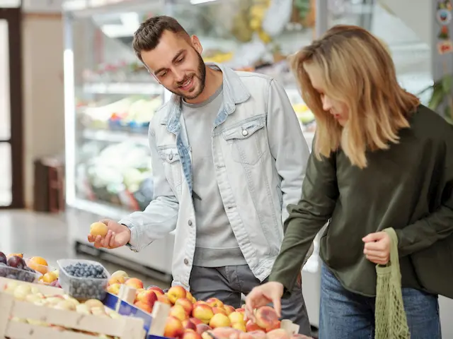 Man and woman shopping for fresh fruit at a grocery store, selecting peaches from a display.