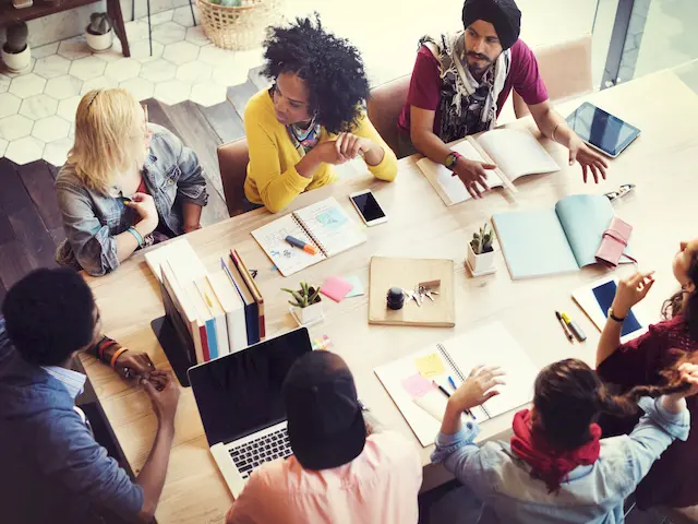 Diverse group of people collaborating around a table with laptops, notebooks, and documents, engaged in a team discussion.