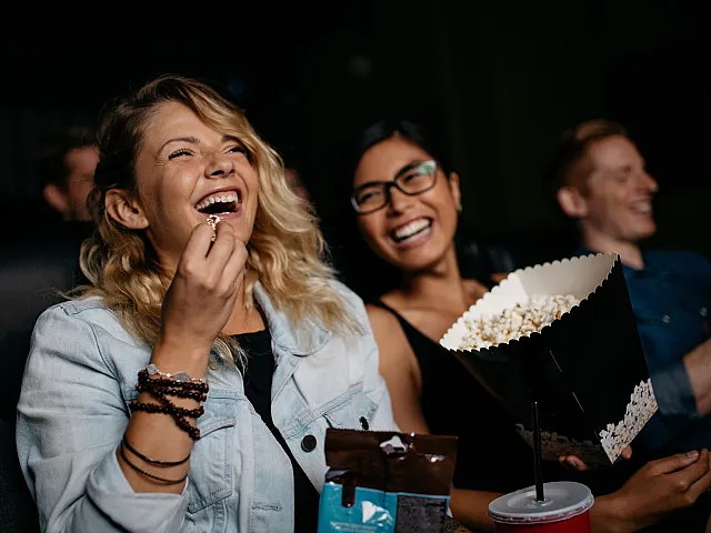 Three people sitting in a movie theater laugh while watching a film, with one person in the foreground holding popcorn and smiling brightly.