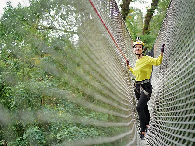 Adventurous woman crossing a thrilling rope bridge, surrounded by lush greenery, wearing a helmet and safety harness for an exciting outdoor experience.