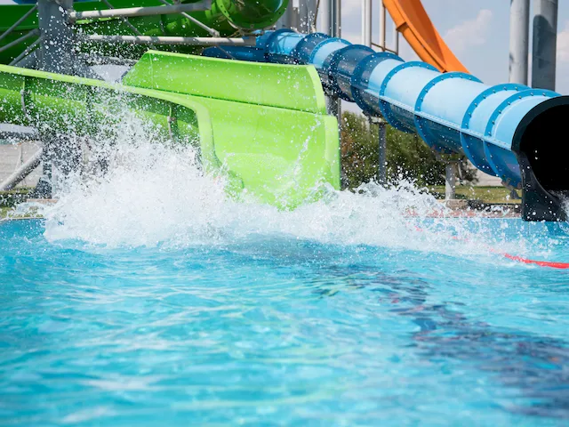 Water slides empty into a bright blue pool, with water splashing as it flows out of green and blue slide tubes on a sunny day.