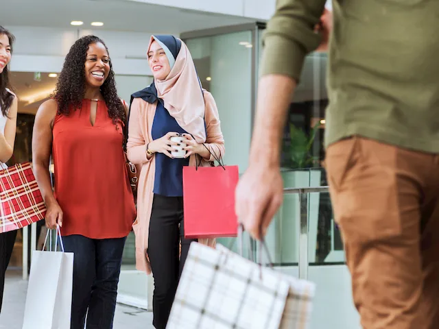 Three women walking together outside a shopping center, smiling and chatting while carrying shopping bags, as another person walks past in the foreground.