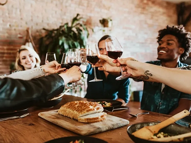 Group of friends gathered around a dining table, raising glasses of red wine in a toast, with food and bread served in a warm, casual setting.