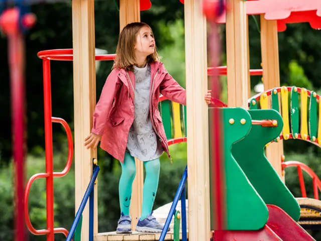 Joyful young girl exploring a colorful playground structure with vibrant green and red elements, surrounded by lush greenery.