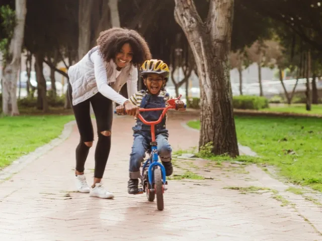 A smiling woman helps a young child ride a bicycle along a paved park path surrounded by trees and greenery. The child is wearing a yellow helmet and laughing while learning to ride, creating a joyful and family-friendly outdoor scene.