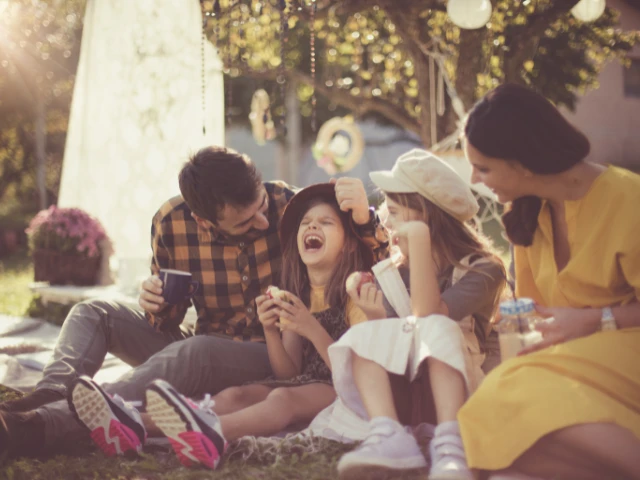 A family of four sits together on a blanket outdoors, laughing and enjoying snacks in a sunlit park setting. Surrounded by trees and warm afternoon light, the group shares a joyful moment near a fountain, creating a welcoming and family-friendly atmosphere.