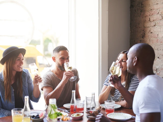 Group of friends gathered around a restaurant table, enjoying food, conversation, and drinks in a bright, casual dining setting.