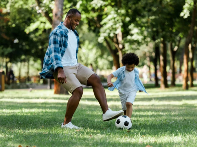 Parent and child playing soccer together in a lush, tree-filled park, enjoying quality time and outdoor recreation on a sunny day.
