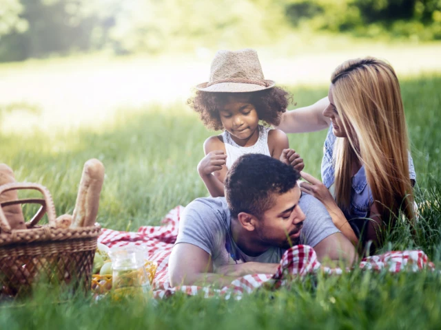 Family enjoying a picnic together in a sunny park, relaxing on a blanket with a picnic basket while spending quality time outdoors in a peaceful, natural setting.