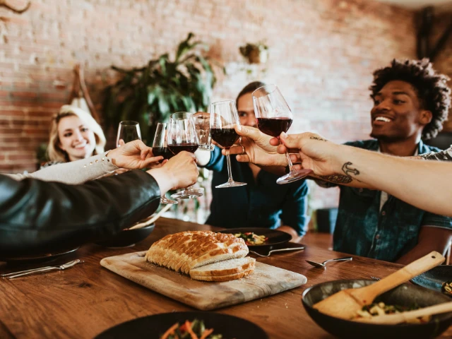 A group of friends gathered around a dining table raising glasses of red wine in a toast, with fresh bread and shared dishes creating a warm, social atmosphere.